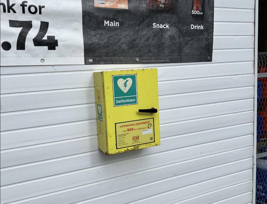 A yellow defibrillator box is mounted on a white wall next to a sign showing drink prices.